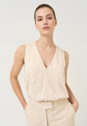 Jeune femme portant un chemisier beige sans manches à col en V et un pantalon taille haute assorti, les mains dans les poches, fond neutre.