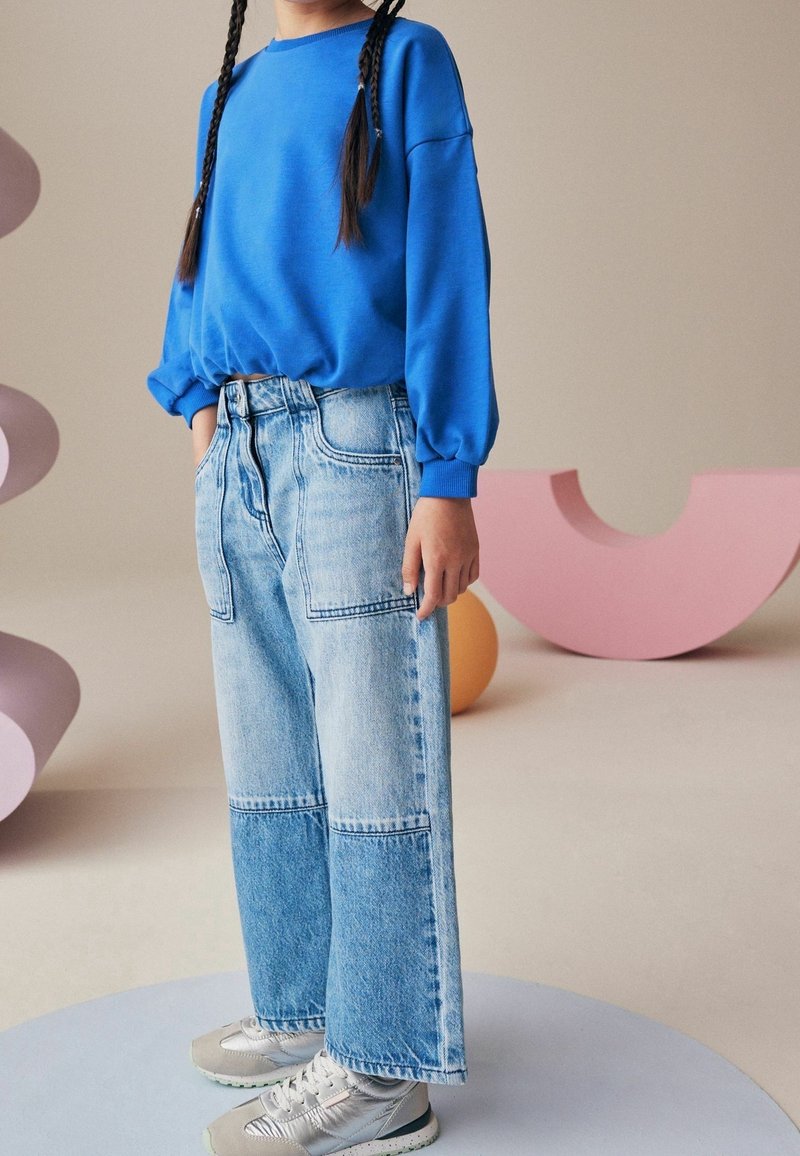 Enfant avec des cheveux tressés portant un sweat-shirt bleu, un jean large bicolore et des baskets argentées, debout sur une plateforme ronde.