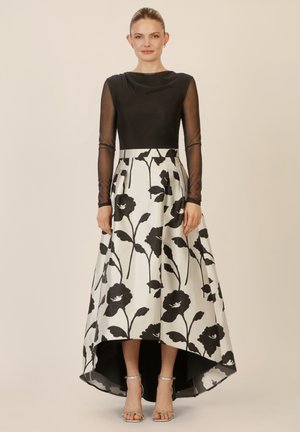 Schwarz tailliertes Oberteil mit transparenten langen Ärmeln, kontrastiert durch einen High-Low-Rock mit großen schwarzen Blumenprints auf einem weißen Hintergrund.
