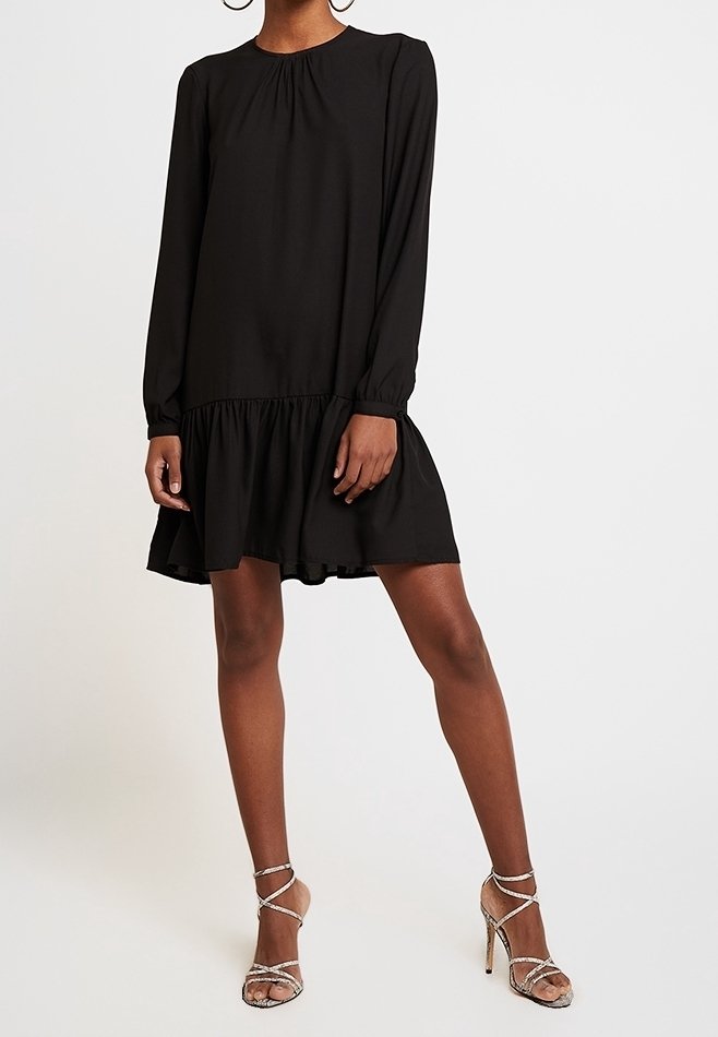 Robe noire à manches longues avec un col froncé et un ourlet volanté, en tissu léger. Le mannequin porte des sandales à talons transparents à brides.