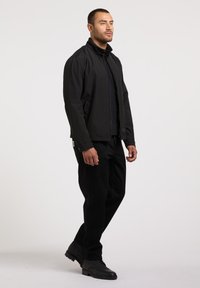 Homme debout portant une veste noire à fermeture éclair, un pantalon noir et des bottes noires, sur un fond clair uni.