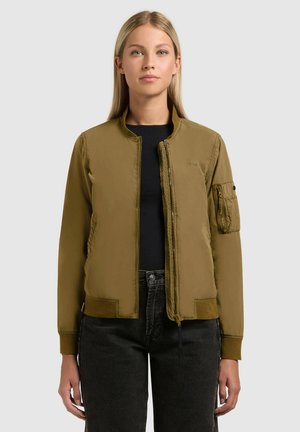 Young woman wearing a khaki bomber jacket over a black shirt and black jeans, standing against a plain light gray background.