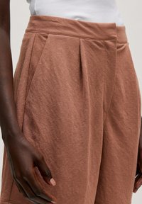 Braune, strukturierte Stoffhose mit hoher Taille, die über vordere Falten und seitliche Taschen verfügt. Glatte Oberfläche mit lockerer Passform.