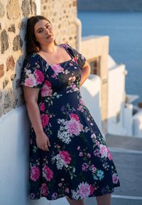 Frau in einem dunklen Blumenkleid lehnt an einer Steinmauer, im Hintergrund weiße Gebäude und das blaue Meer bei Sonnenuntergang.