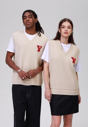 Gilets en tricot beige avec des encolures en V, ornés de patchs rouges brodés en forme de 'Y'. Portés par-dessus des t-shirts blancs, associés à un jean noir et une jupe en cuir.