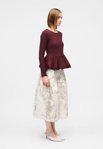 Bluză maro cu mâneci lungi și tiv cu volane, asortată cu o fustă evazată albă cu model floral. Model purtând pantofi cu toc ascuțiți albi.