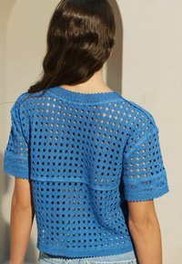 Modrý pletený top s háčkováním, s diamantovými výřezy, krátkými rukávy a vykrojeným lemem. Zadní strana ukazuje vrstvený design.