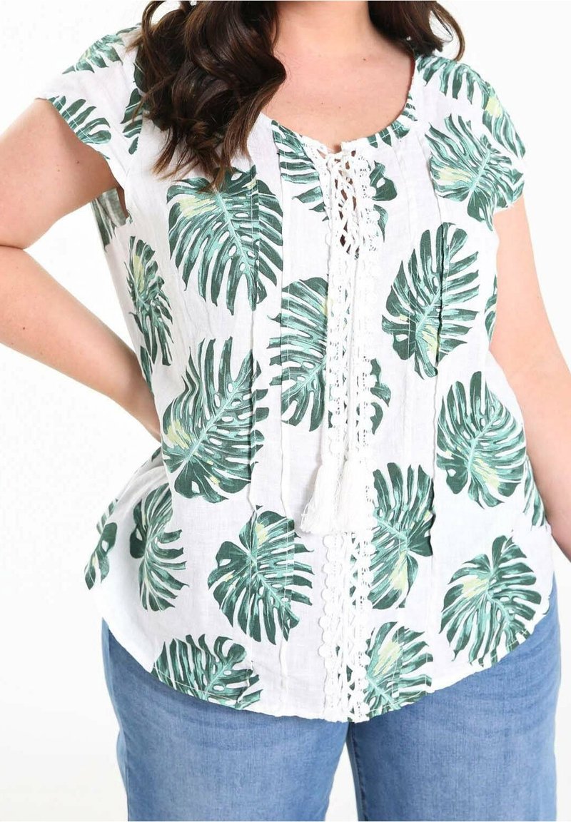 Femme portant un chemisier blanc à manches courtes avec imprimé feuilles tropicales vertes et laçage à l'avant, associé à un jean bleu.