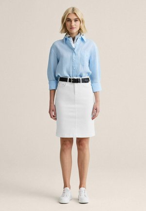 Kvinde stående iført en lyseblå skjorte med knapper, hvid knælængde nederdel med sort bælte og hvide sneakers mod en neutral baggrund.