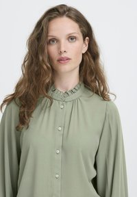 Young woman with wavy brown hair wearing a light green buttoned blouse with a ruffled collar, facing forward against a plain background.