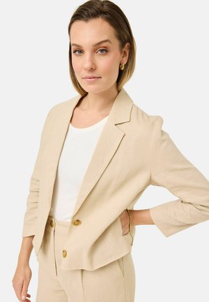 Frau mit kurzen Haaren, die einen beigen Blazer und eine Hose über einem weißen Hemd trägt, posiert mit einer Hand in der Hüfte vor einfachem Hintergrund.