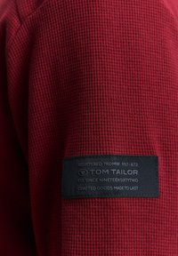 Tessuto rosso testurizzato con un motivo a nido d'ape e un'etichetta nera. L'etichetta presenta del testo bianco con il nome del marchio e la data di fondazione per Tom Tailor.