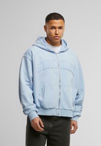 Lys blå zip-up hættetrøje lavet af blødt stof med broderet logo, der har en frontlomme og en afslappet pasform.
