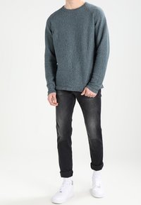 Suéter de punto texturizado en azul verdoso con cuello redondo, combinado con unos pantalones oscuros enrollados y zapatillas blancas. Tejido suave y diseño casual.