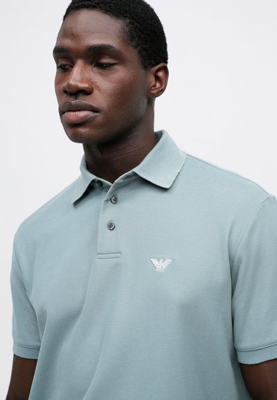 Ανοιχτό μπλε polo μπλουζάκι κατασκευασμένο από υφασμάτινη υφή, διαθέτοντας πλάκα με τρία κουμπιά και μικρό κεντητό λογότυπο στο αριστερό στήθος.