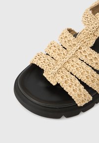 Brązowo-czarna sandałka z plecionymi beżowymi paskami, zaokrąglonym noskiem i grubą czarną podeszwą z teksturowanymi krawędziami.