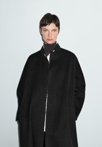 Persona con capelli corti e scuri, che indossa un cappotto oversize nero e un dolcevita scuro, in piedi su uno sfondo chiaro e uniforme.