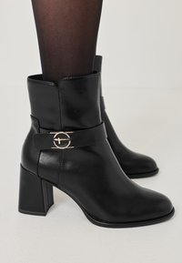 Bottines en cuir noir avec un bout carré, un talon bloc de hauteur intermédiaire, et une sangle décorative avec un accent circulaire en ton or.