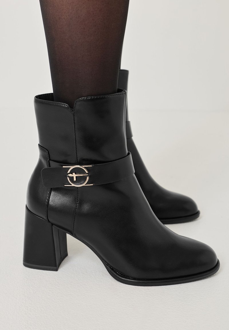 Bottines en cuir noir avec un bout carré, un talon bloc de hauteur intermédiaire, et une sangle décorative avec un accent circulaire en ton or.