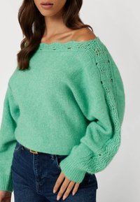 Maglione lavorato a maglia verde menta con design scollato sulle spalle, caratterizzato da dettagli intricati nella lavorazione lungo le maniche e i polsini.