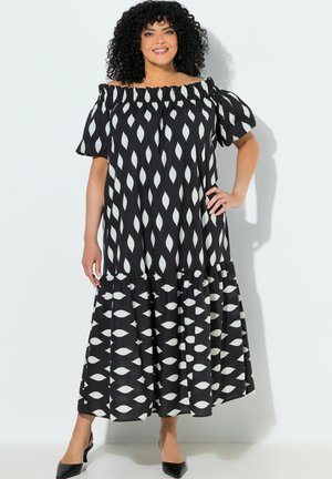 Kobieta o kręconych włosach, ubrana w czarną sukienkę z odkrytymi ramionami i białymi liściastymi wzorami, stoi z jedną ręką na biodrze i uśmiecha się.