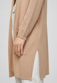 Ljusbeige cardigan med texturerad stickning, sidoklipp och framfickor, bärs över vita byxor.
