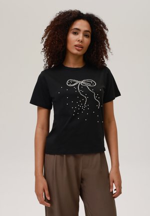 Camiseta negra de manga corta con un diseño de lazo adornado con perlas y perlas esparcidas. Tela suave, corte informal.