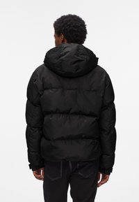 Háttal látható személy fekete, bélelt kapucnis pufi dzsekit és sötét nadrágot visel, fehér háttér előtt.