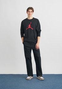 Zwarte sweater met lange mouwen en een rood Jumpman-logo, gecombineerd met een zwarte broek en sneakers met grijze en gele accenten.