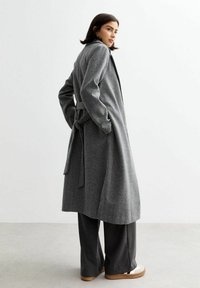 Manteau à chevrons gris avec une taille ceinturée, manches longues et col, associé à un pantalon large gris et des chaussures crème.