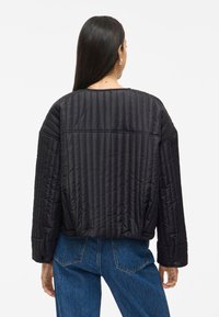 Juodas quiltinis švarkas su laisvu siluetu, apvaliu kaklu, ilgomis rankovėmis ir matomomis siūlėmis. Pora su mėlynomis džinsomis.