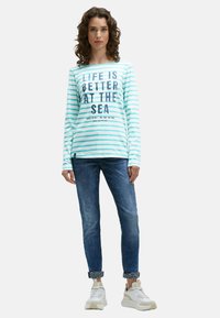 Junge Frau mit lockigem Haar, die ein mintgrünes, gestreiftes Langarmshirt mit maritimem Aufdruck, hochgekrempelte Jeans und weiße Sneakers trägt und steht.