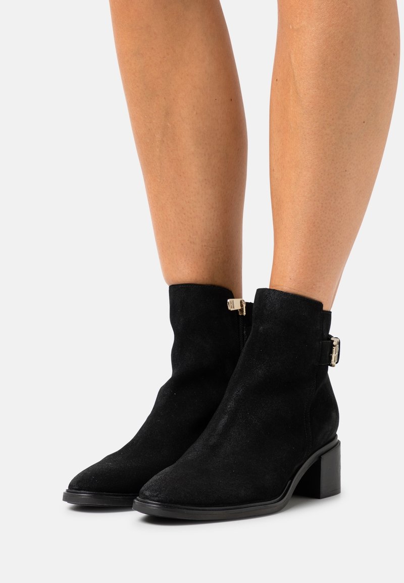 Bottines en daim noir avec fermeture éclair latérale et détail de boucle dorée, portées sur jambes nues avec un petit talon bloc et un bout pointu.
