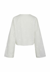 Blouse blanche à manches longues, coupe courte, avec un col rond et un motif floral ajouré détaillé sur le tissu.