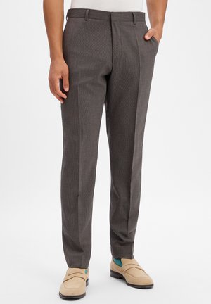 Man draagt op maat gemaakte donkergrijze nette broek met zijzakken, beige loafers en teal sokken, staand tegen een witte achtergrond.
