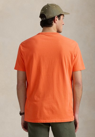 Narancssárga rövid ujjú pólót, olívazöld nadrágot és olívazöld sapkát viselő ember hátulnézetből, egyszínű háttér előtt.