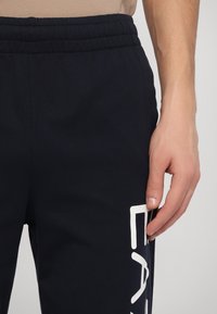 Mørkeblå joggebukser med elastisk midje, med et fremtredende hvitt logodesign på venstre side og en glatt stofftekstur.