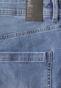 Poche arrière d'un jean en denim bleu clair avec des coutures visibles et un patch de marque en cuir noir portant l'inscription "DENIM STREET ONE".