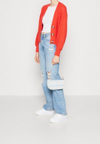 Red knitted cardigan with three buttons, paired with a white cropped top and distressed light blue flared jeans. White sneakers and a blue plaid handbag.