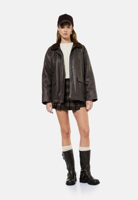 Giacca di pelle marrone con colletto di pelliccia, maglione bianco lavorato a maglia, gonna a quadri e stivali marroni al ginocchio con fibbie e calze color crema.