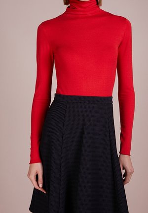 Femme portant un col roulé moulant rouge à manches longues, rentré dans une jupe noire texturée taille haute à silhouette évasée.
