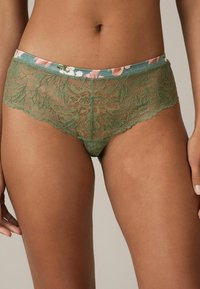 Olīvzaļš mežģīņu bikini ar ziedu raksta jostas daļu, kurai ir sarežģītas mežģīņu detaļas un viļņainas malas, pievienojot papildus tekstūru.