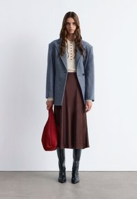 Grauer Wollblazer über einem cremefarbenen Strickoberteil und einem braunen satinierten Rock, kombiniert mit schwarzen ledernen kniehohen Stiefeln und einer roten Wildledertasche.