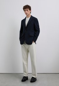 Joven de pie con las manos en los bolsillos, vistiendo un blazer azul marino, una camisa blanca, pantalones crema y zapatos negros, frente a un fondo simple.