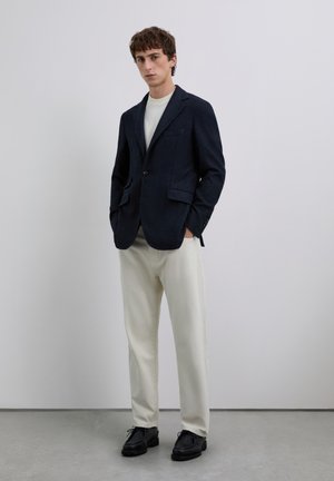 Joven de pie con las manos en los bolsillos, vistiendo un blazer azul marino, una camisa blanca, pantalones crema y zapatos negros, frente a un fondo simple.