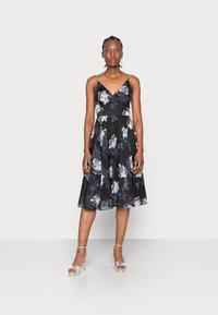 Vestido floral em preto com rosas brancas e azuis, apresentando um corpete justo, alças ajustáveis e uma saia rodada. Sapatos de salto prateados complementam o visual.