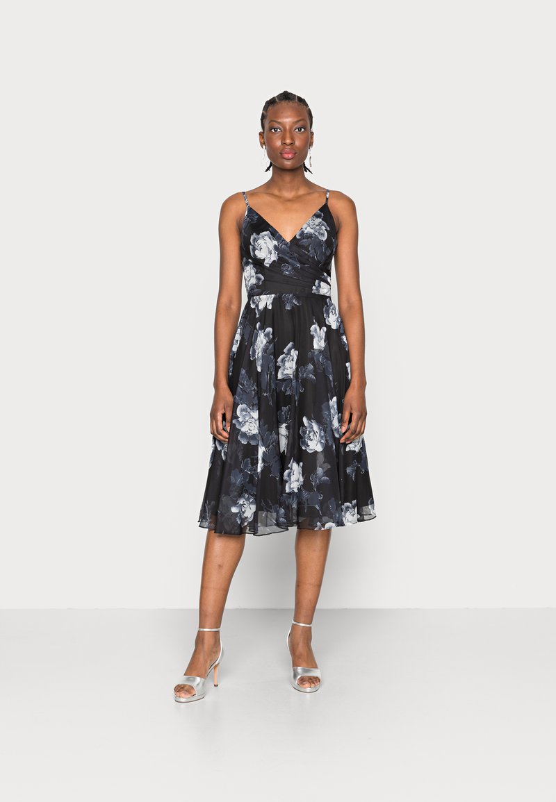 Vestido floral em preto com rosas brancas e azuis, apresentando um corpete justo, alças ajustáveis e uma saia rodada. Sapatos de salto prateados complementam o visual.