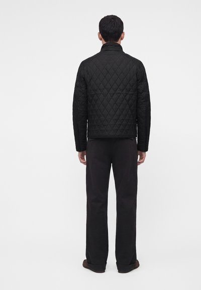 Homme portant une veste matelassée noire et un pantalon noir, se tenant de dos devant un arrière-plan clair uni.