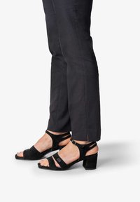 Mustat mokkapintaiset sandaalit, joissa on leveät nauhat, avonaiset varpaat ja paksu kantapää. Yhdistetty tummaan housuuttuihin, joissa on halkio helmassa, korostaen kenkämallia.