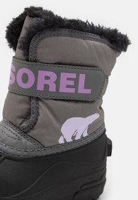 Botín gris de invierno con ribete de piel sintética negra, suela de goma y un logo "SOREL" en color púrpura. Presenta un gráfico de oso en un lado.